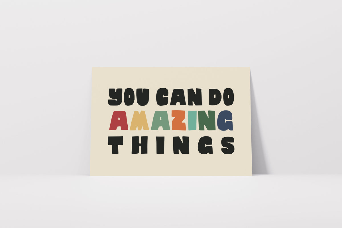 A square cream-colored art print or card with the motivational quote 'YOU CAN DO AMAZING THINGS' in bold black letters, with 'AMAZING' in multicolor block letters, leaning against a white wall on a white floor.